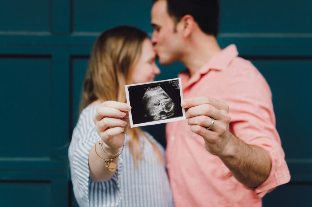 Couple kissing each other holding a baby scan picture up to the camera